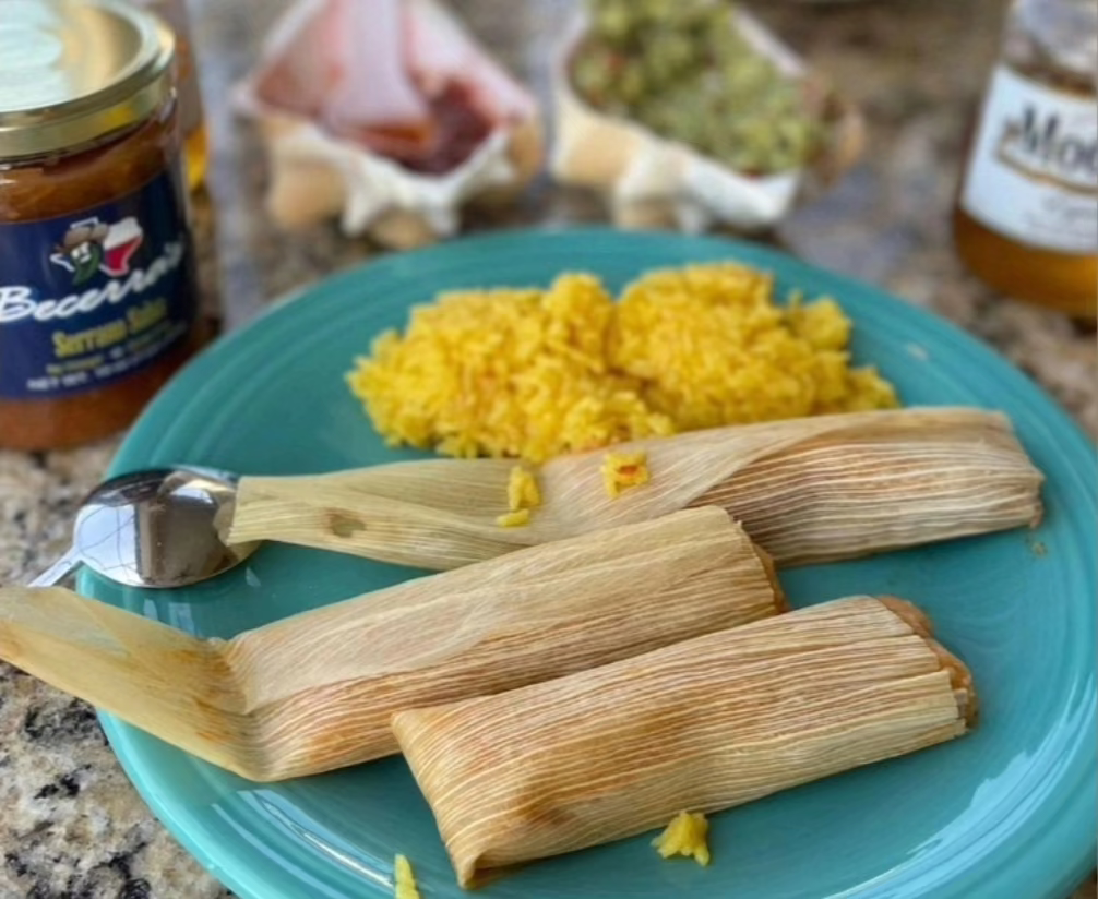 Three Becerra Brand Tamales on a blue plate with yellow rice, surrounded by Becerra Brand Serrano Salsa on a countertop.
